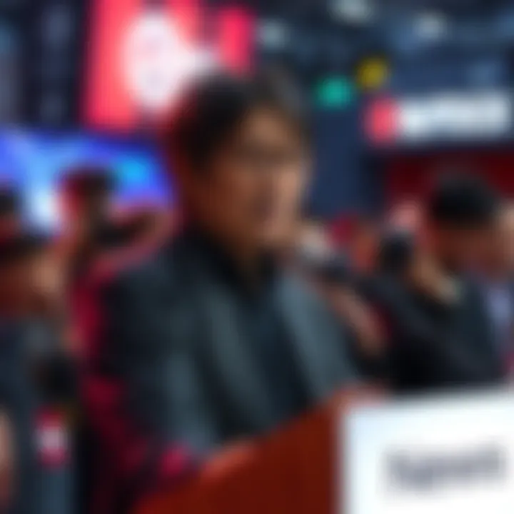Hideo Kojima stands at a podium, speaking into a microphone with a thoughtful expression, surrounded by gaming fans and media cameras.
